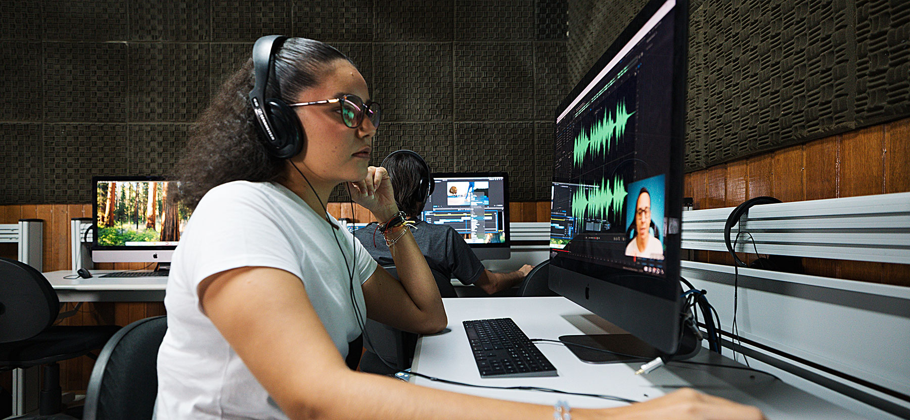 Mulher de óculos editando vídeo em estúdio de edição de áudio e vídeo com computadores e equipamento de alta qualidade, foco em pós-produção de mídia digital.